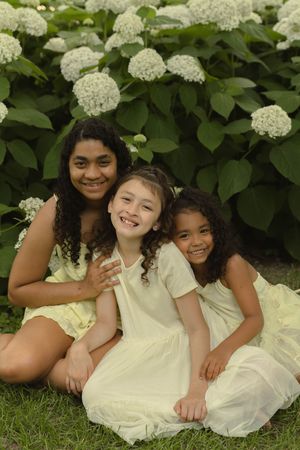 Three children in cream-colored dresses pose together among blooming white hydrangea bushes.