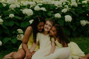 Children in light summer dresses share laughs while posing near white hydrangea flowers.