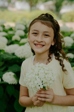 Child in white dress smiles while holding fresh-picked hydrangea flowers.