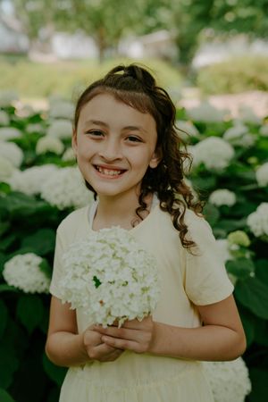 Young child in white dress holds white hydrangea flowers while smiling in garden setting.