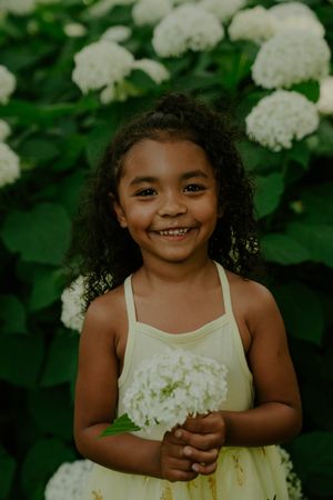 Child in cream dress poses playfully with white hydrangea blooms in garden.