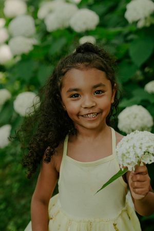 Child in light dress holds white hydrangea flowers against green garden background.