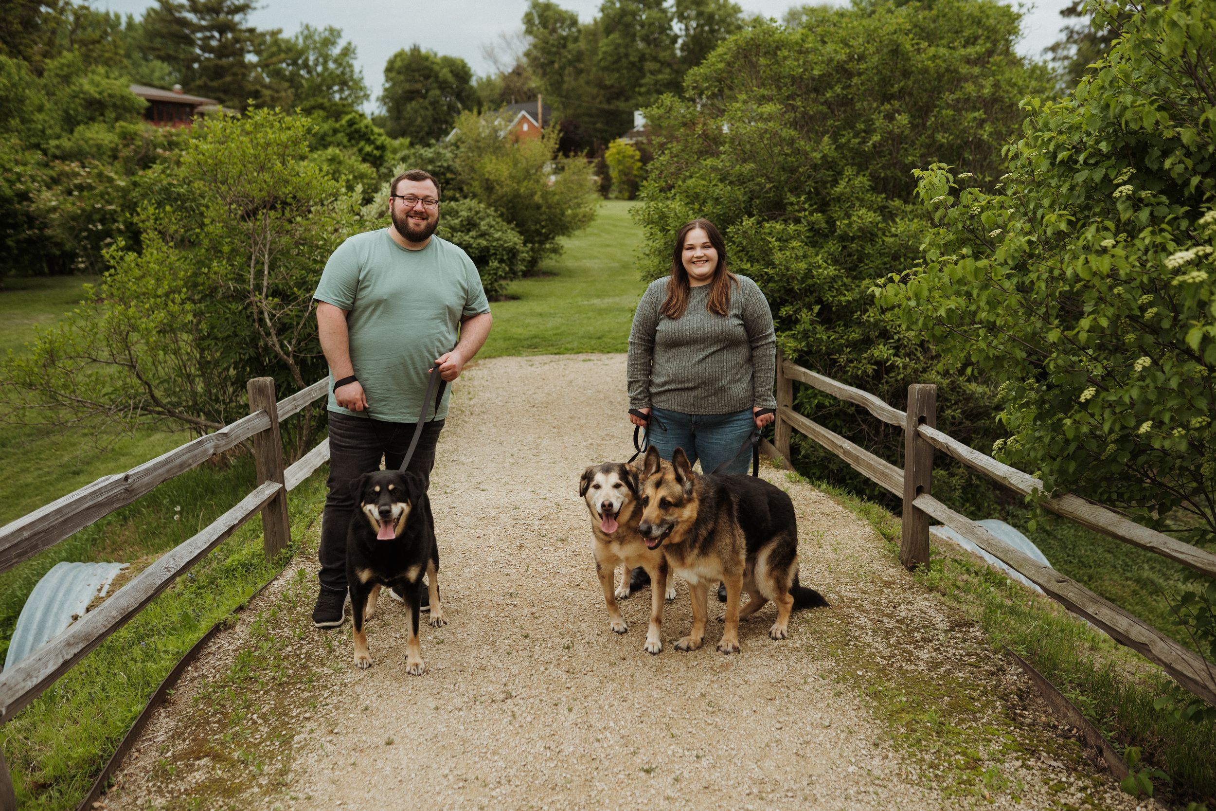A couple walks their dogs along a gravel path in a peaceful park setting.