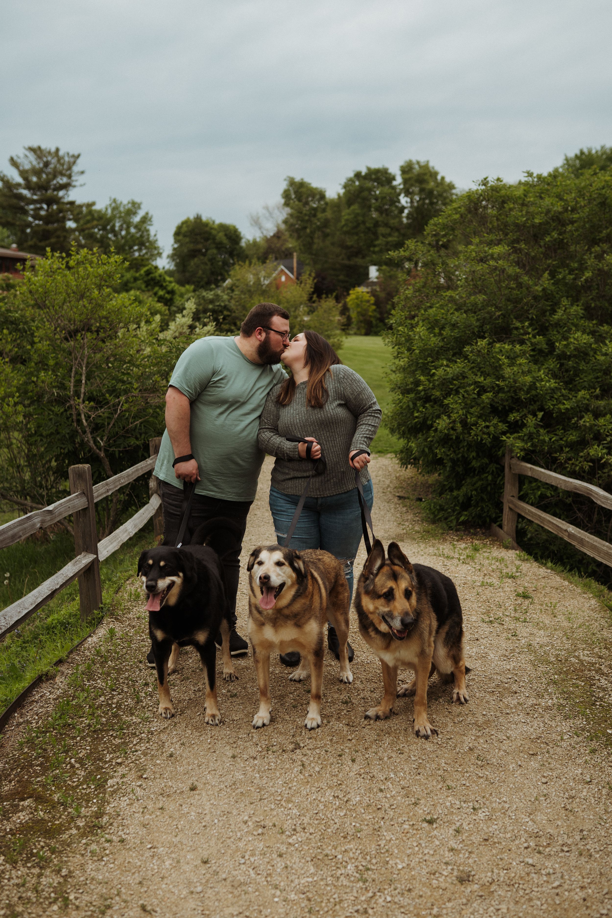 A couple poses with their four dogs on a gravel trail surrounded by greenery.