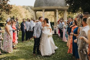 A joyous outdoor garden wedding ceremony with guests gathered around a domed gazebo on a sunny afternoon.
