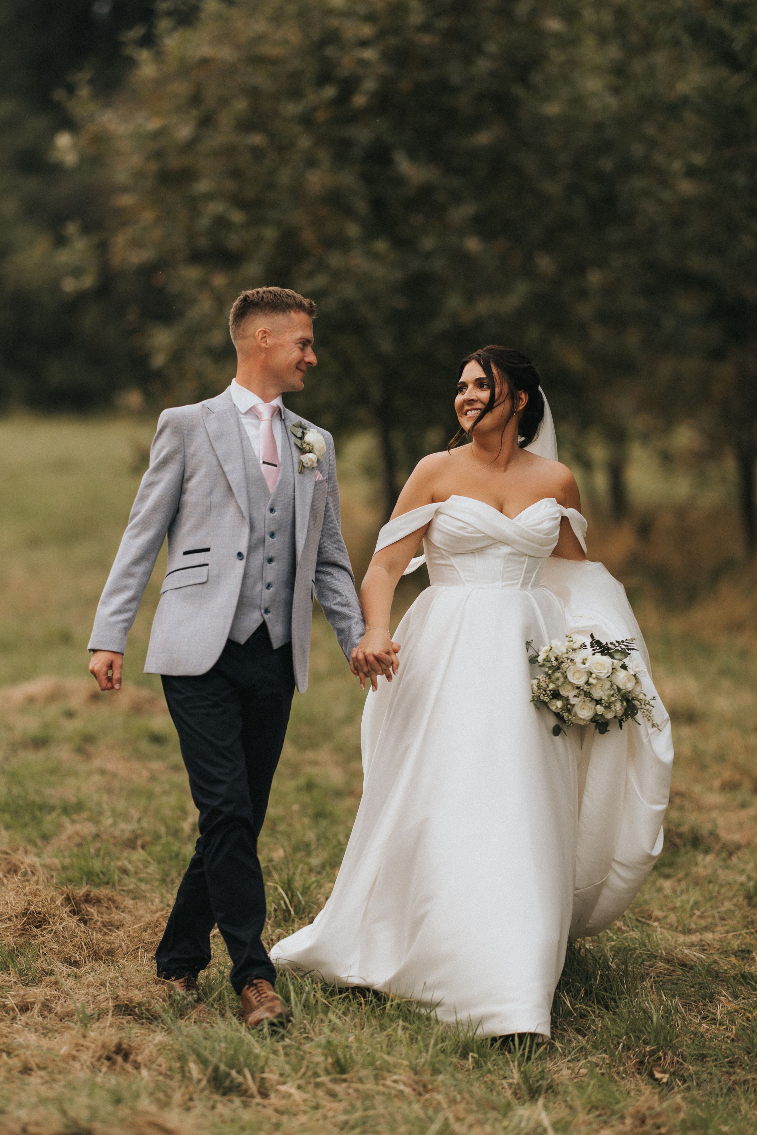 Newlyweds walk hand in hand through a grassy field, bride in white gown holding bouquet and groom in light gray suit.