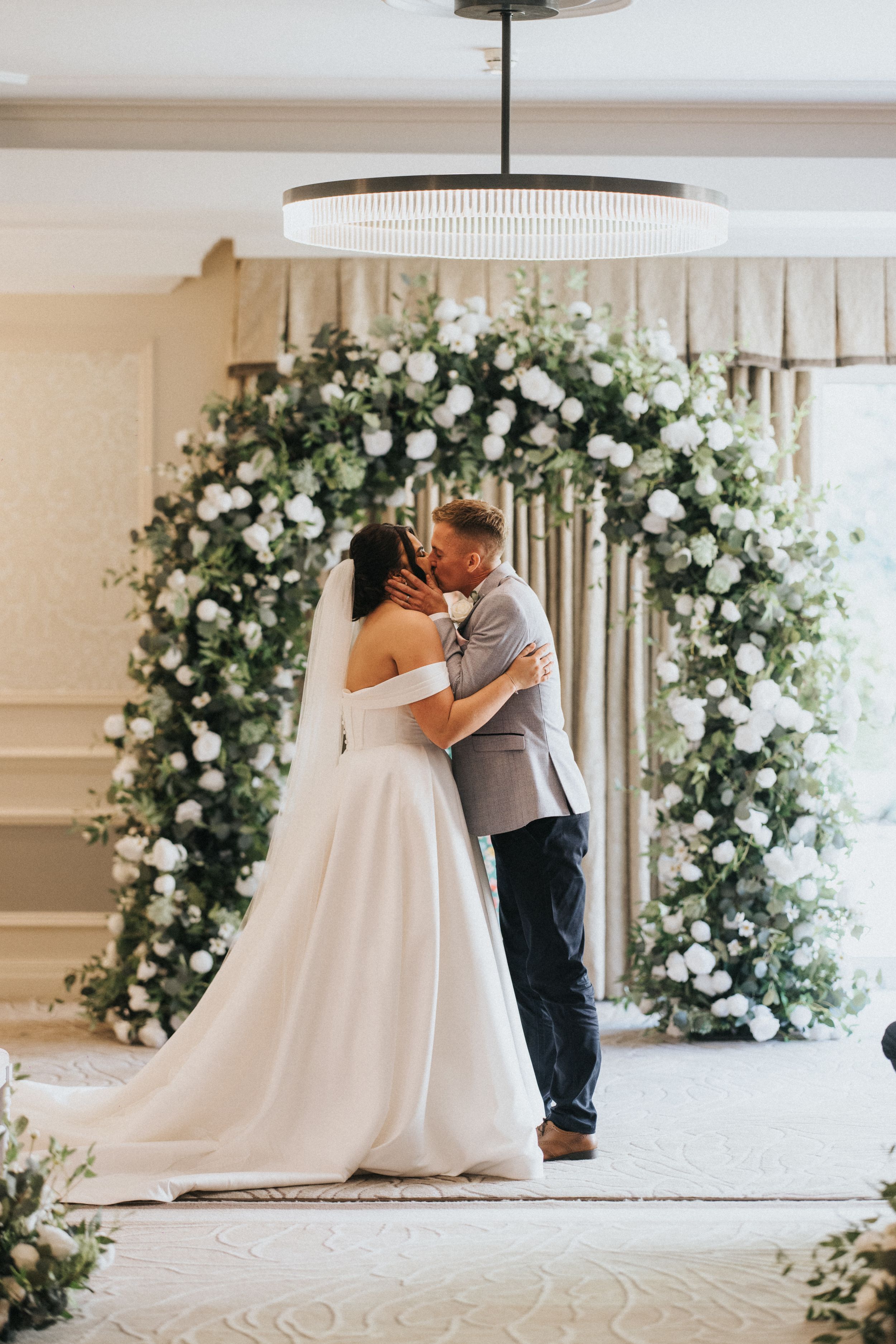 Couple shares romantic wedding moment under elegant white floral arch with roses and greenery at indoor venue.