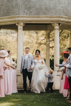 Wedding party gathers under an elegant stone gazebo in a garden setting with bridesmaids in pink dresses.