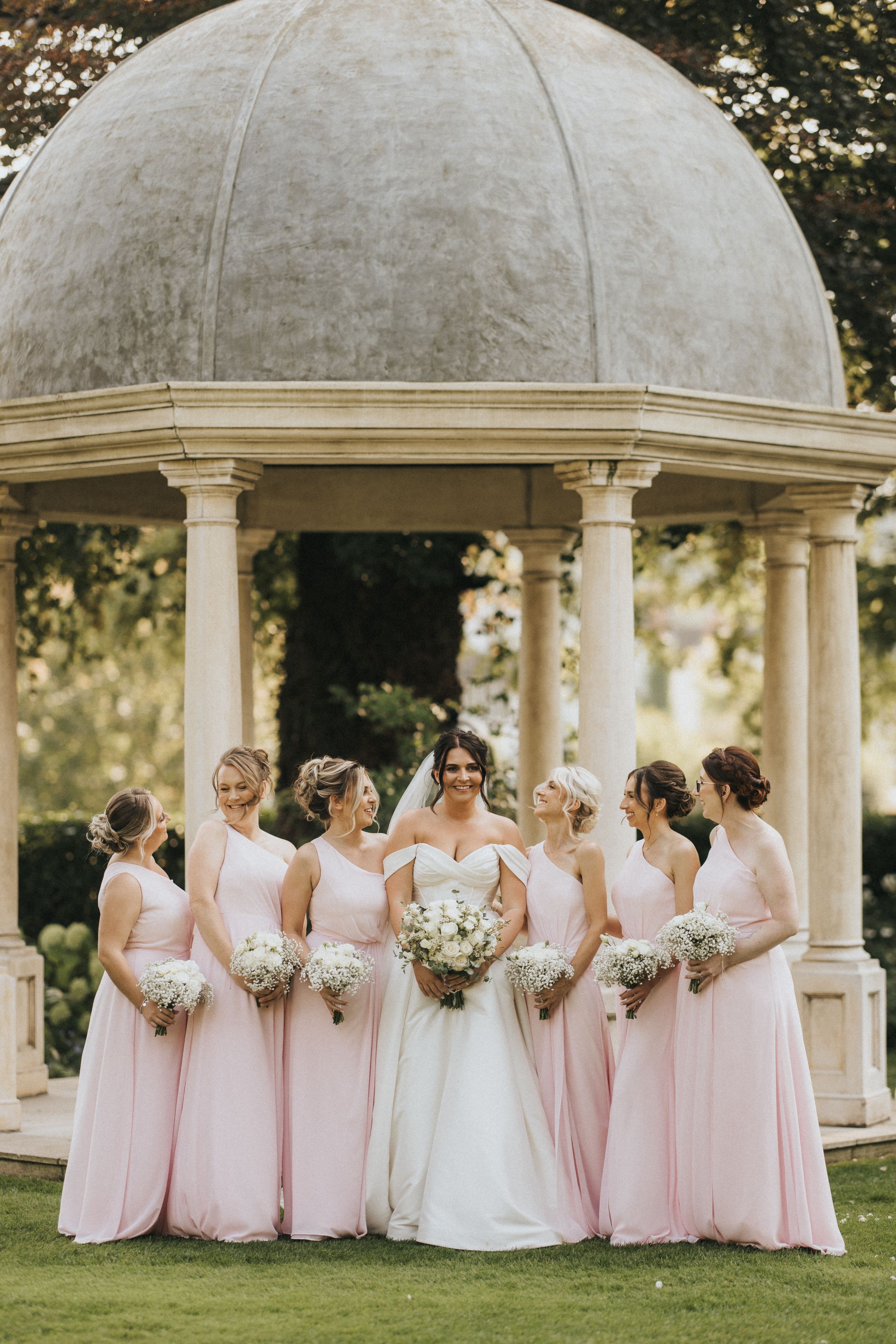 Wedding party members in pink dresses and a white gown pose together under an elegant stone gazebo in a garden setting.