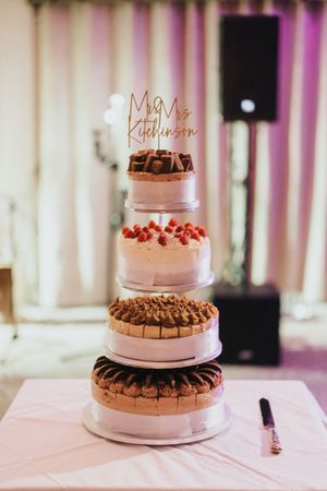 Elegant five-tier wedding cake with pink frosting, chocolate decorations, and strawberries topped with a gold 'Mr & Mrs' sign.