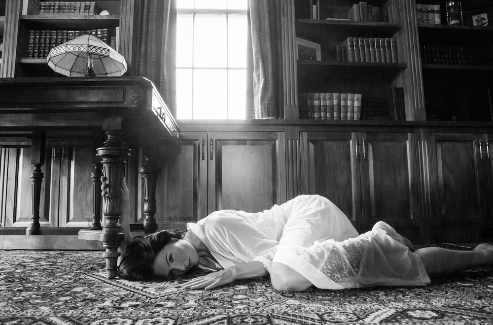 A figure in a white dress lies on a vintage library floor beside wooden paneled walls and bookshelves.