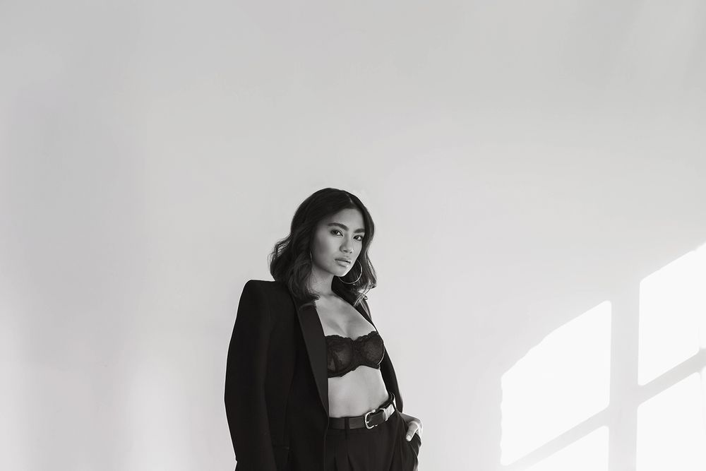 A stylish figure poses dramatically against a wall in black clothing with dramatic lighting and shadows.