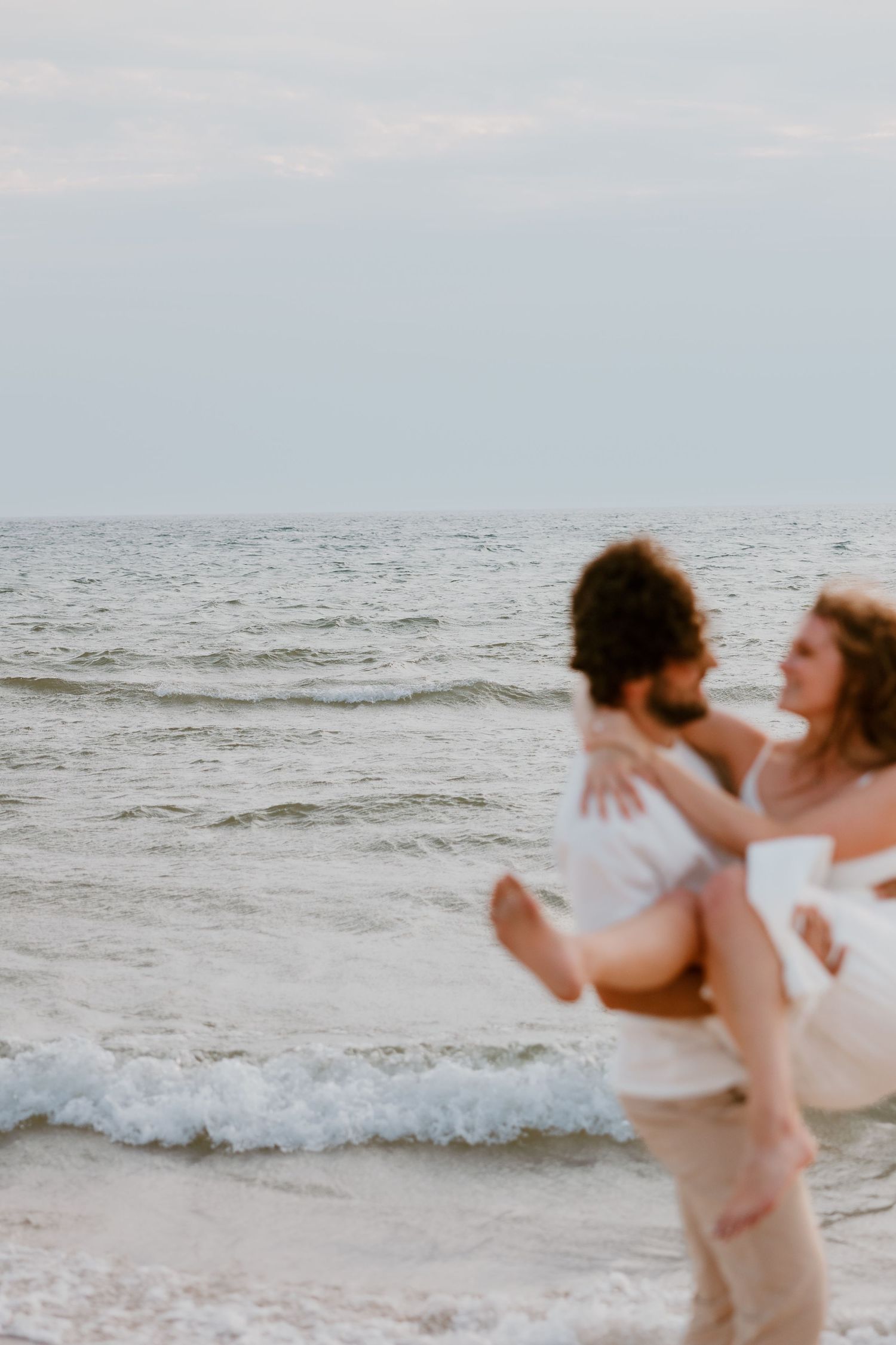 Couple embracing on a beach with waves crashing behind them in a romantic beach photoshoot sequence. Novi Engagement Wedding Photographer