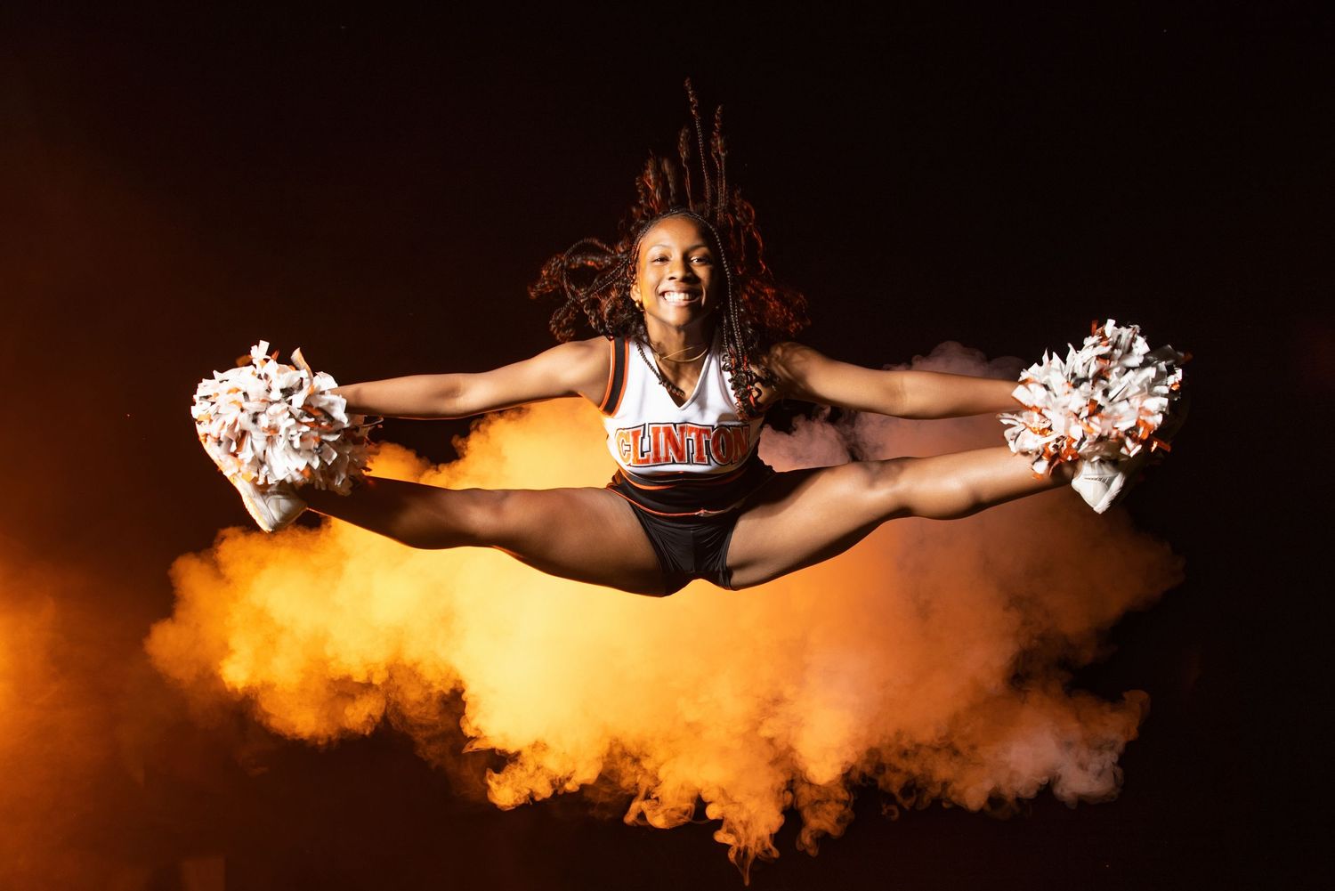 A cheerleader in a black and white uniform performs an aerial split jump with pom-poms against dramatic orange smoke.