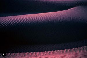Desert sand dunes illuminated in purple and pink hues creating mesmerizing ripple patterns at twilight.