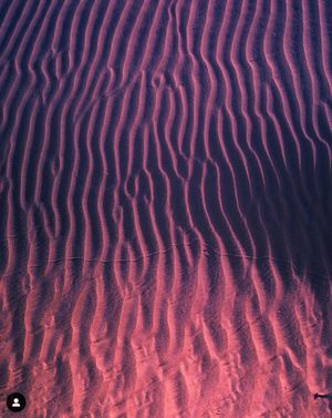 Abstract pattern of wavy purple and red ripples in sand creating mesmerizing natural texture and shadows.