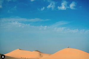 Smooth orange sand dunes rise against a bright blue sky with wispy white clouds in the desert landscape.