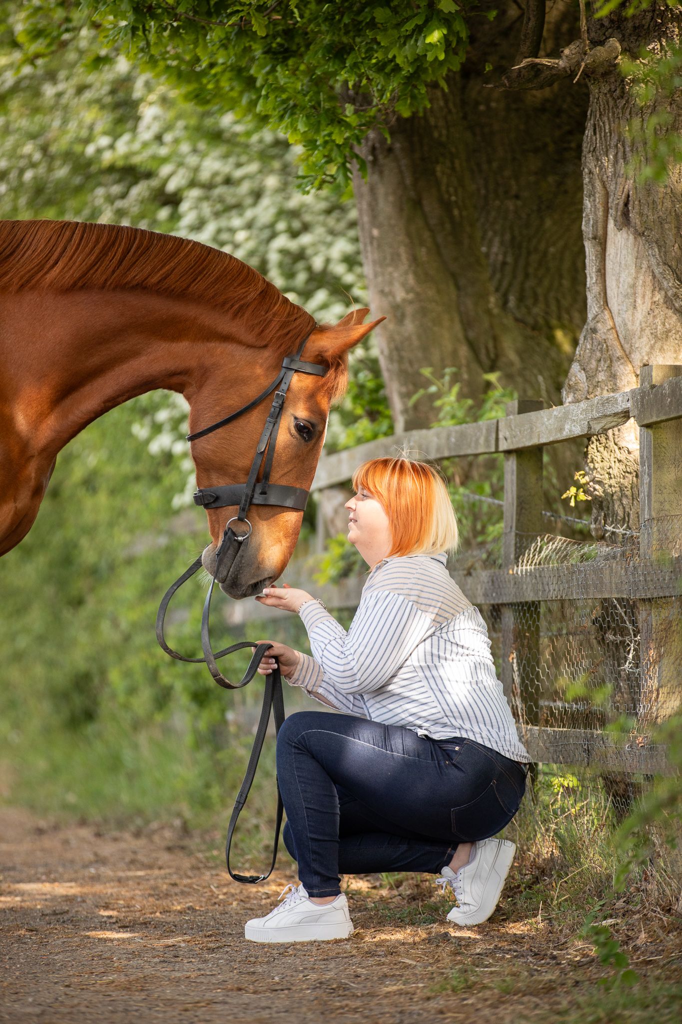 A brown horse and someone stand together in a forest clearing with purple wildflowers blooming around them.