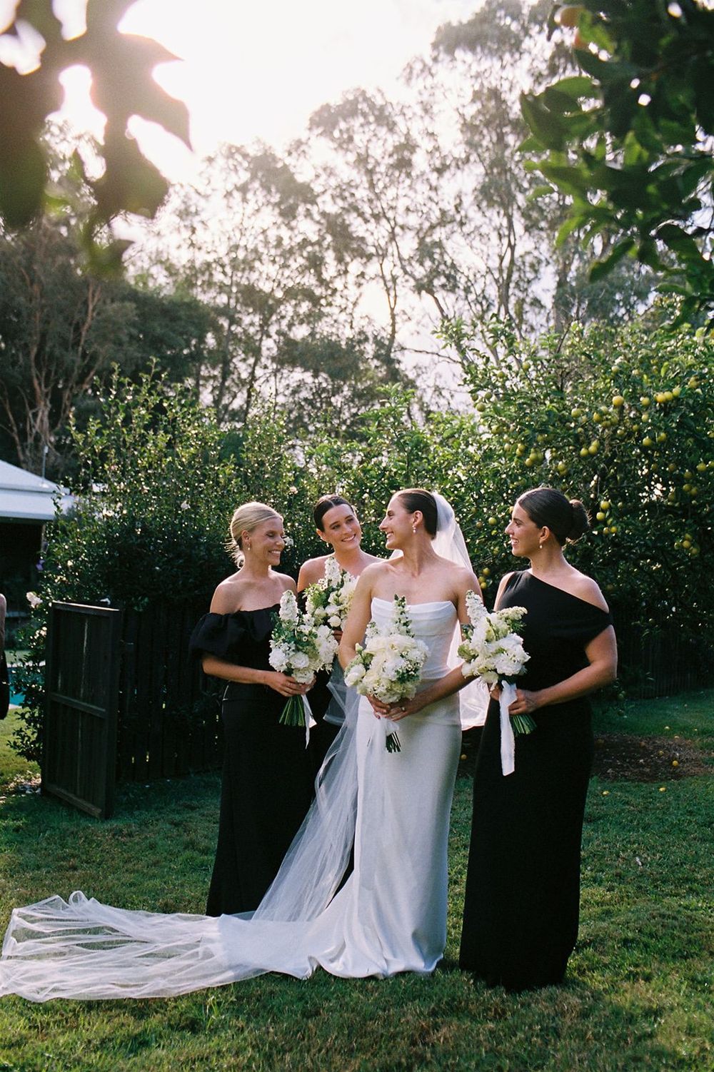 Bride in white gown with bridesmaids in black off-shoulder dresses holding white bouquets in garden setting.
