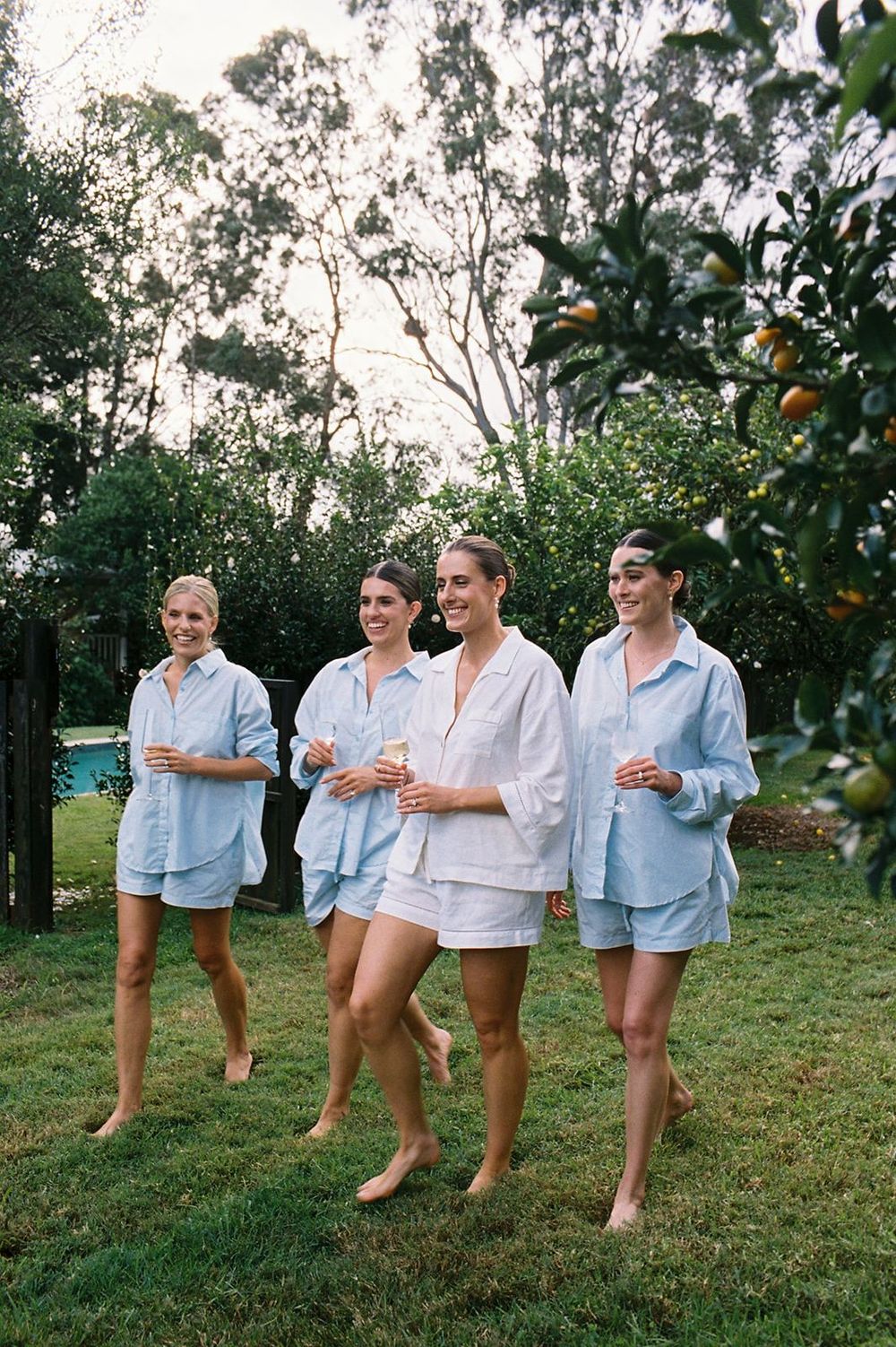 Four women in matching blue and white pajama sets walk barefoot on grass near a pool and orange tree.