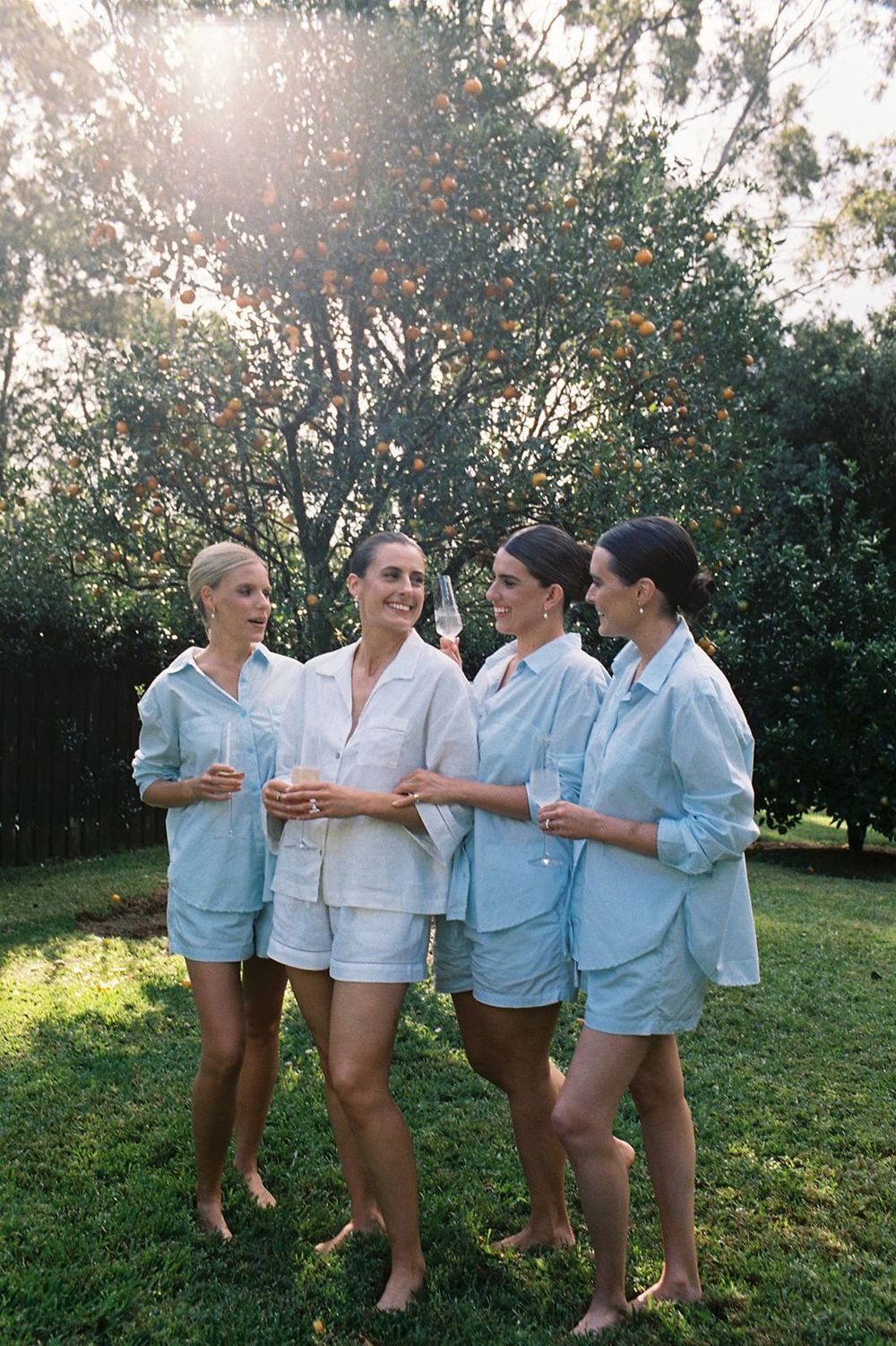 Four women in matching light blue pajama sets laughing together outdoors on green grass near orange trees.