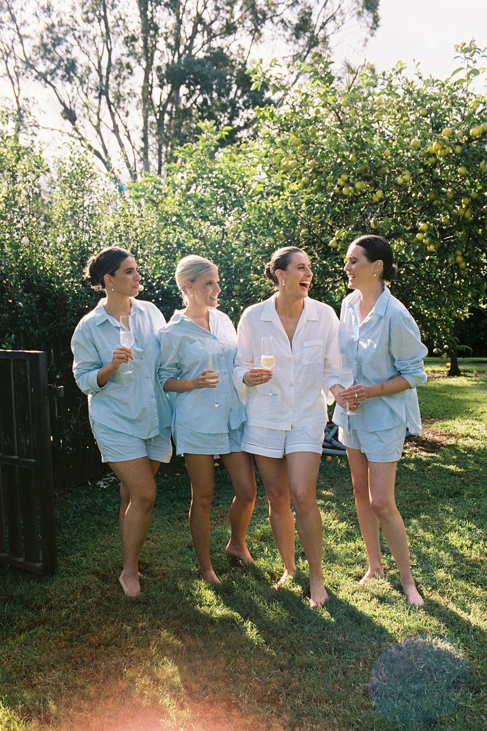 Four women in matching pajamas laughing and holding champagne glasses outdoors on a sunny green lawn.