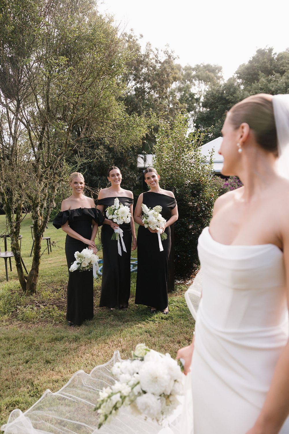Wedding party in black dresses stands outdoors among trees during a sunny celebration.
