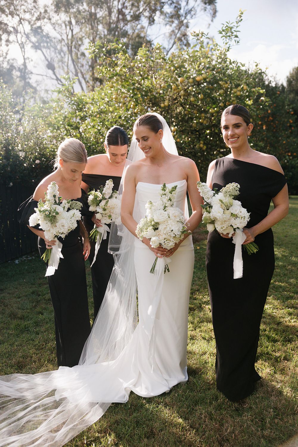 Group photo of bridesmaids in black dresses holding white floral bouquets in a garden setting.