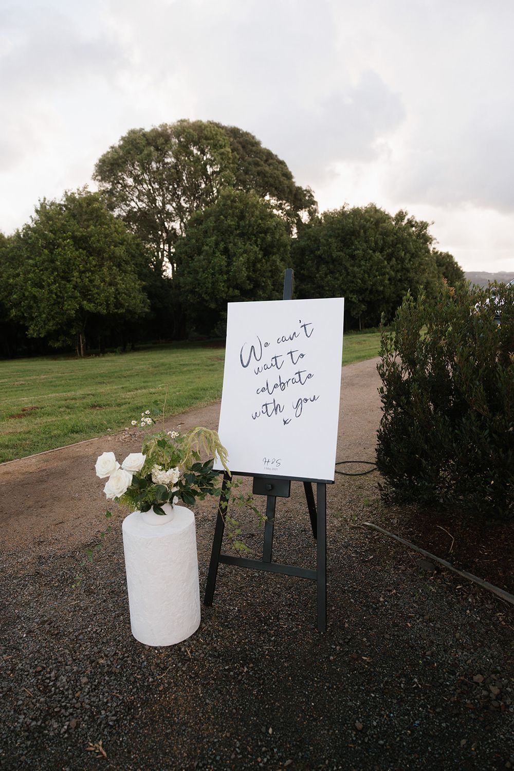 A welcome sign on an easel stands next to white flowers in a vase on a path with trees in the background.
