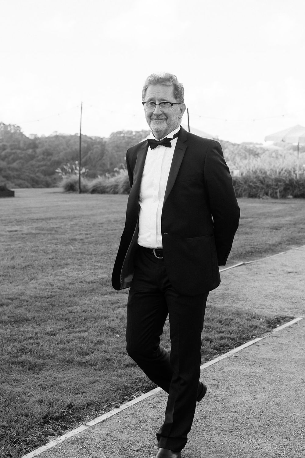 Black and white portrait of a person in a formal tuxedo and bowtie standing on a pathway outdoors.