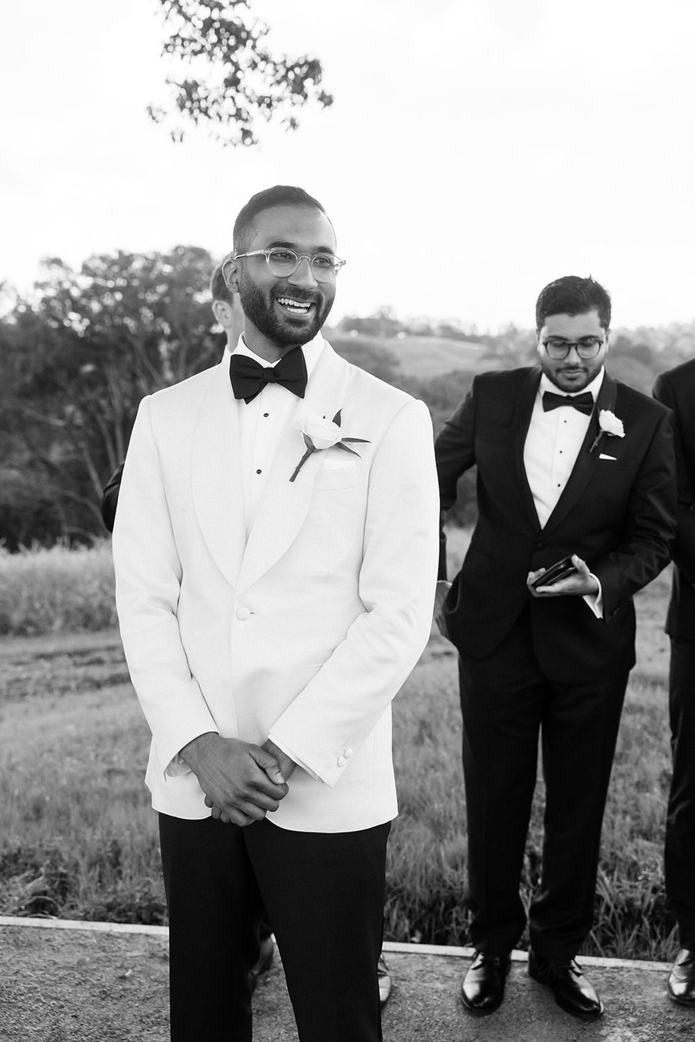 Black and white wedding photo showing two well-dressed men in tuxedos and bow ties standing outdoors.