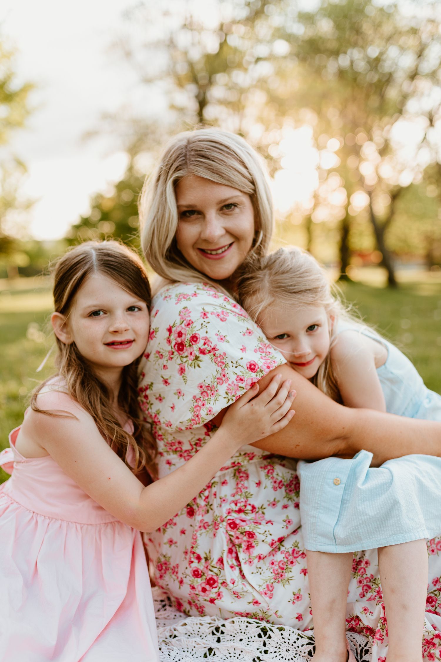 a mom in a flower dress hugging her two daughters, one sitting in her lap and the other behind her hugging her