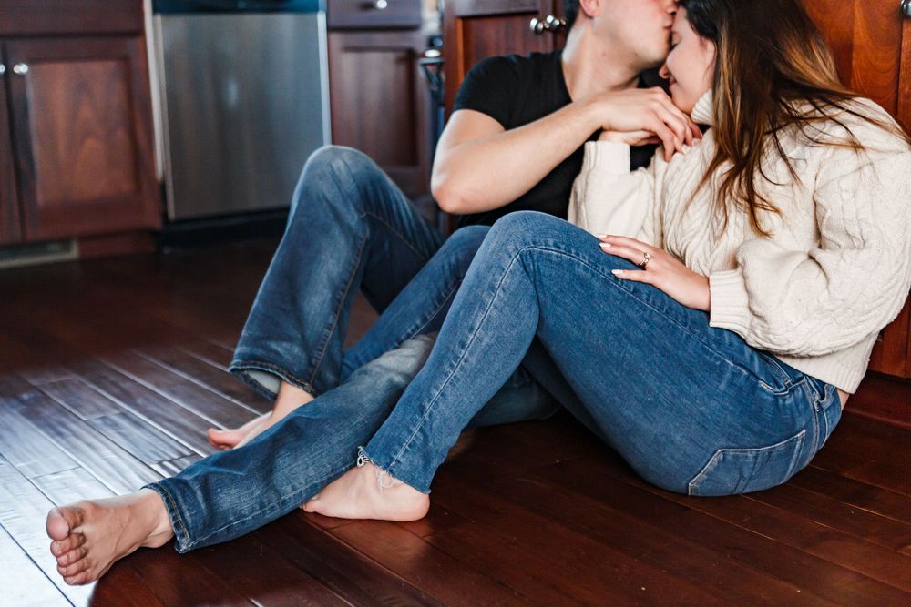 Couple wearing jeans and a white sweater sit together on dark hardwood flooring in intimate poses.