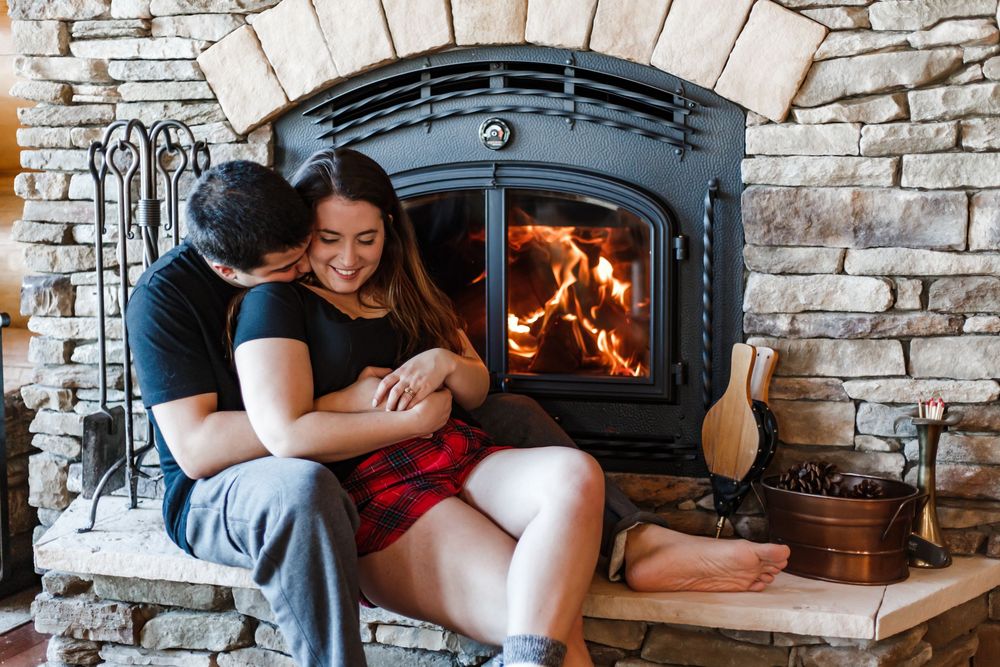 A couple cuddles by a cozy stone fireplace with glowing flames and firewood on a winter evening.
