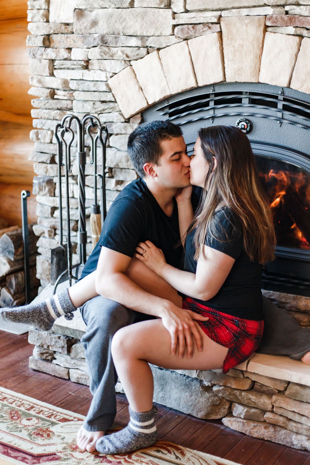 A romantic couple sits intimately by a rustic stone fireplace in a cozy indoor setting.