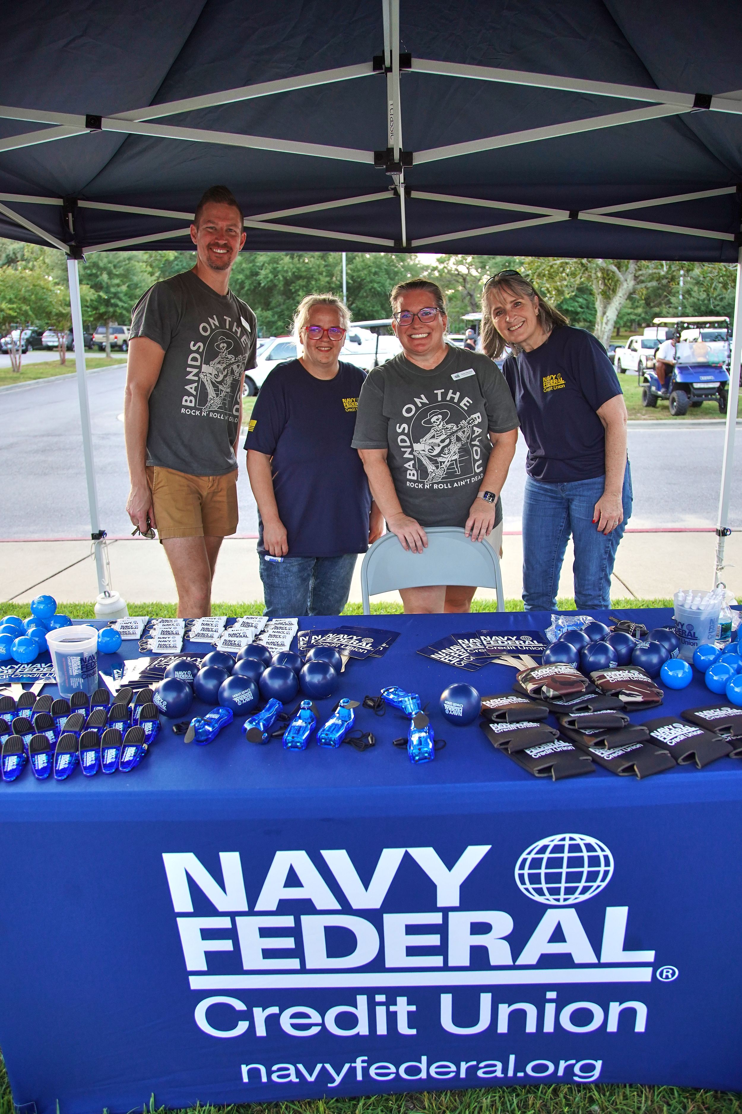 Four Navy Federal Credit Union volunteers smile at a branded booth displaying promotional items at an outdoor event.