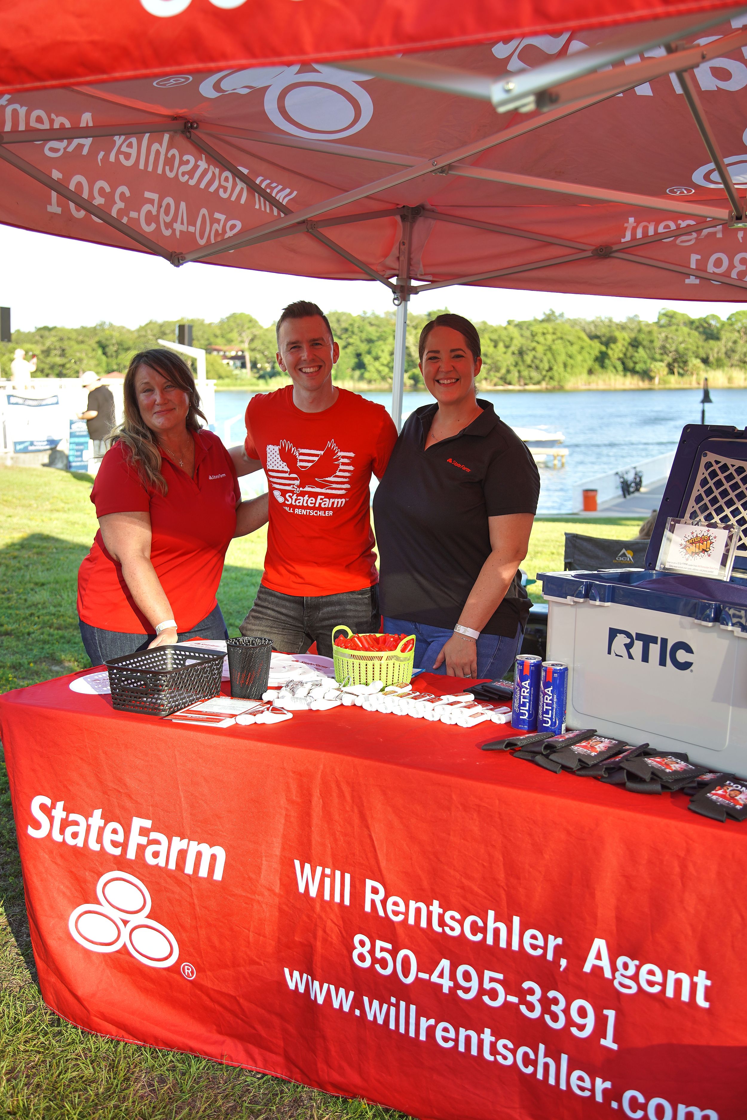 Three smiling staff at a State Farm booth for Will Rentschler, Agent, at an outdoor event near a lake.