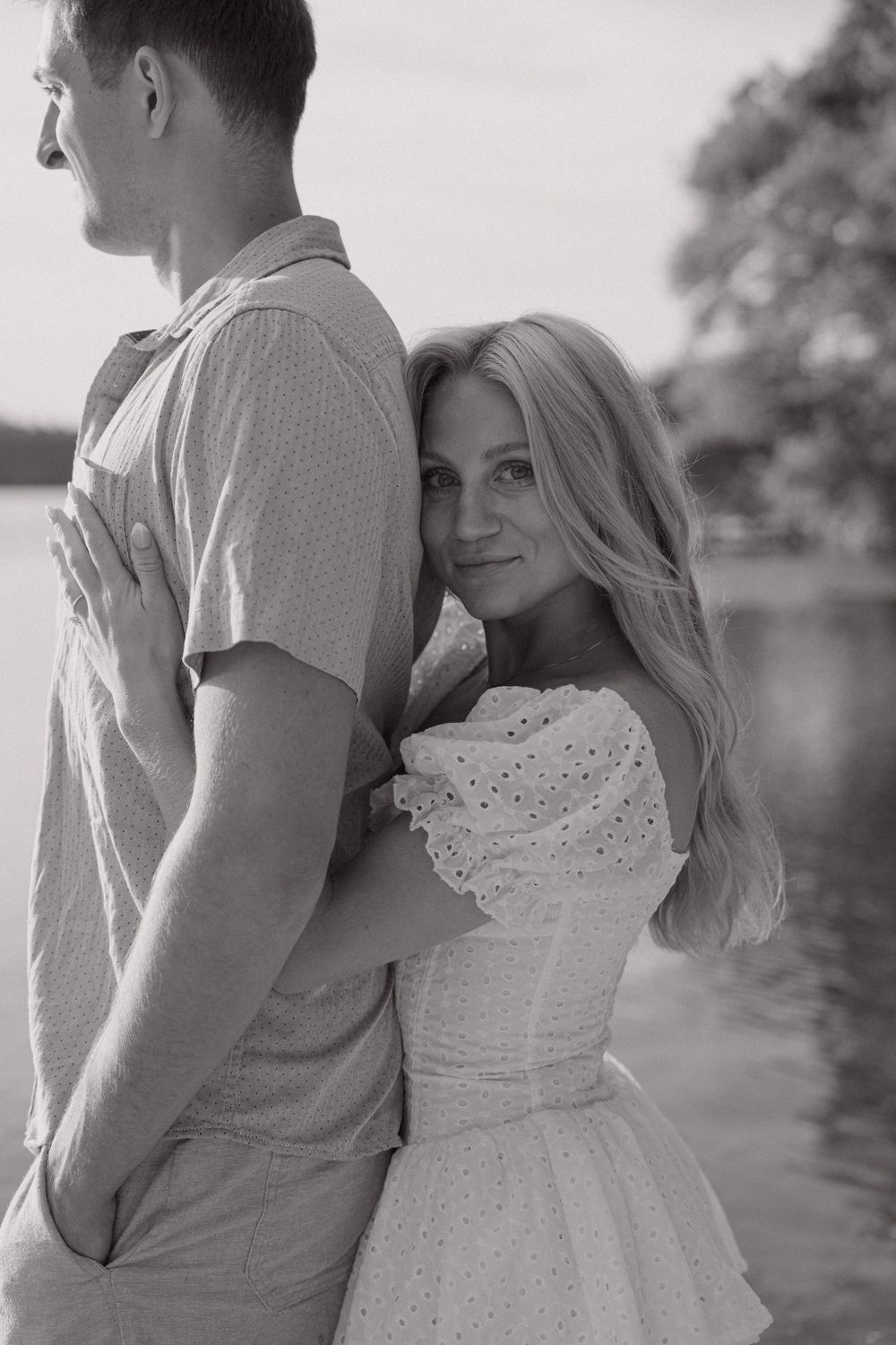 A romantic black and white portrait of a couple by a lake, with her wearing a white dress.