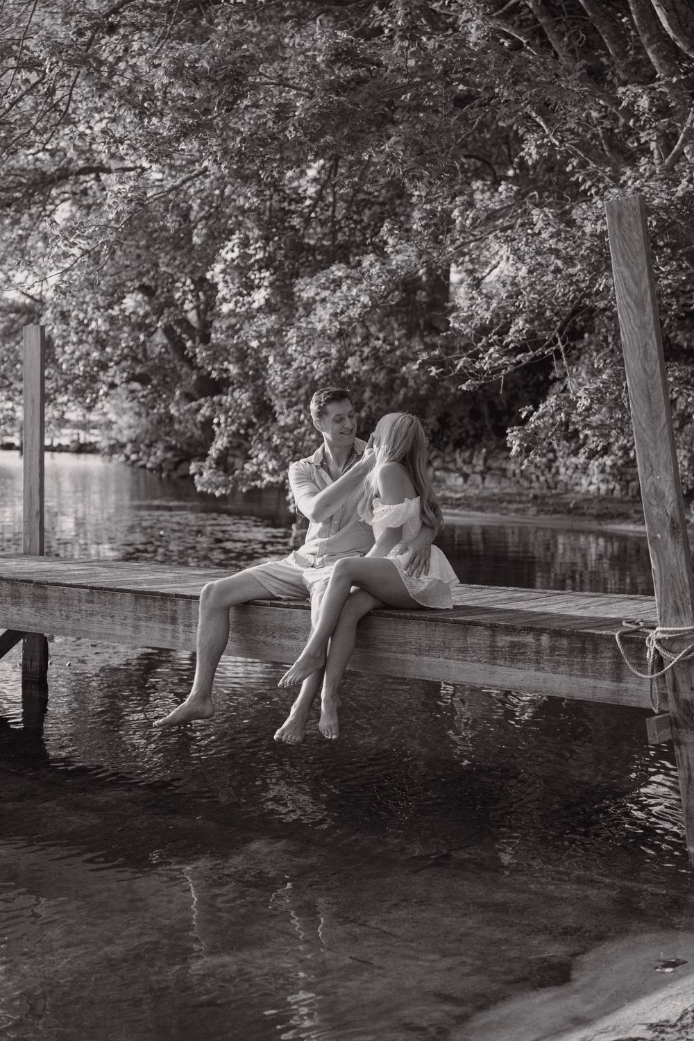 A romantic black and white photo of a couple sitting on a dock over calm water.