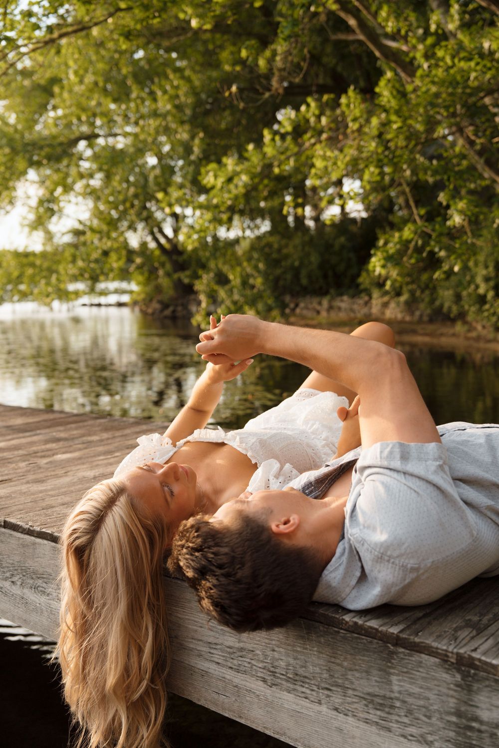 Romantic couple lying on wooden dock by lake with green trees in background during golden hour.