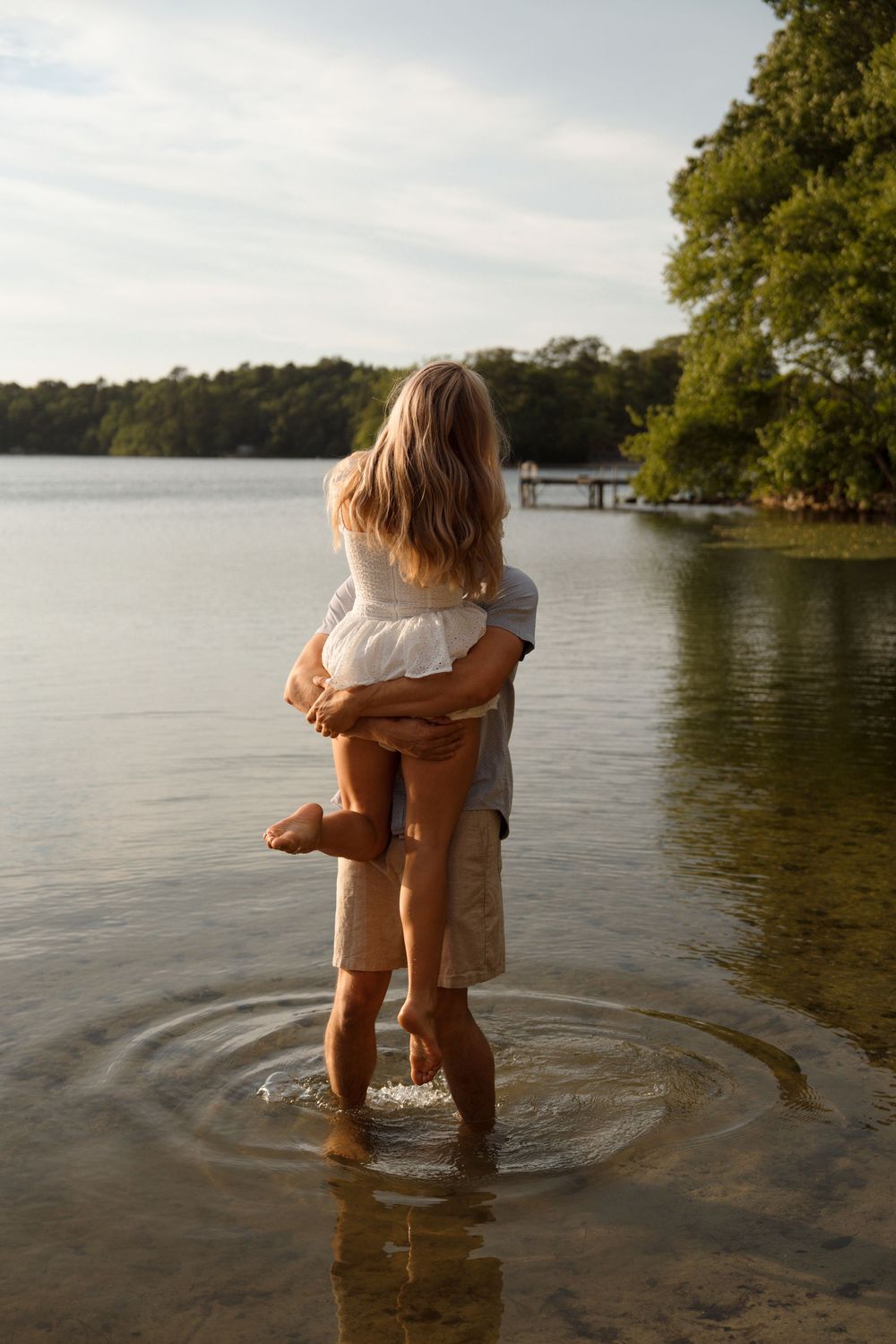 Romantic couple embraces while standing in shallow lake water at sunset with trees and dock in background.