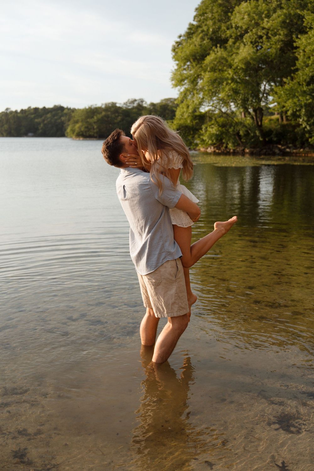 A romantic couple embraces while standing in shallow lake water during golden hour with trees in background.