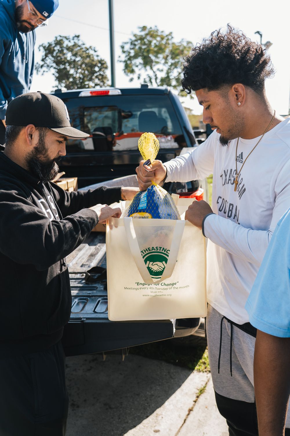 People exchanging items from paper bags in an outdoor setting with cars visible in the background.
