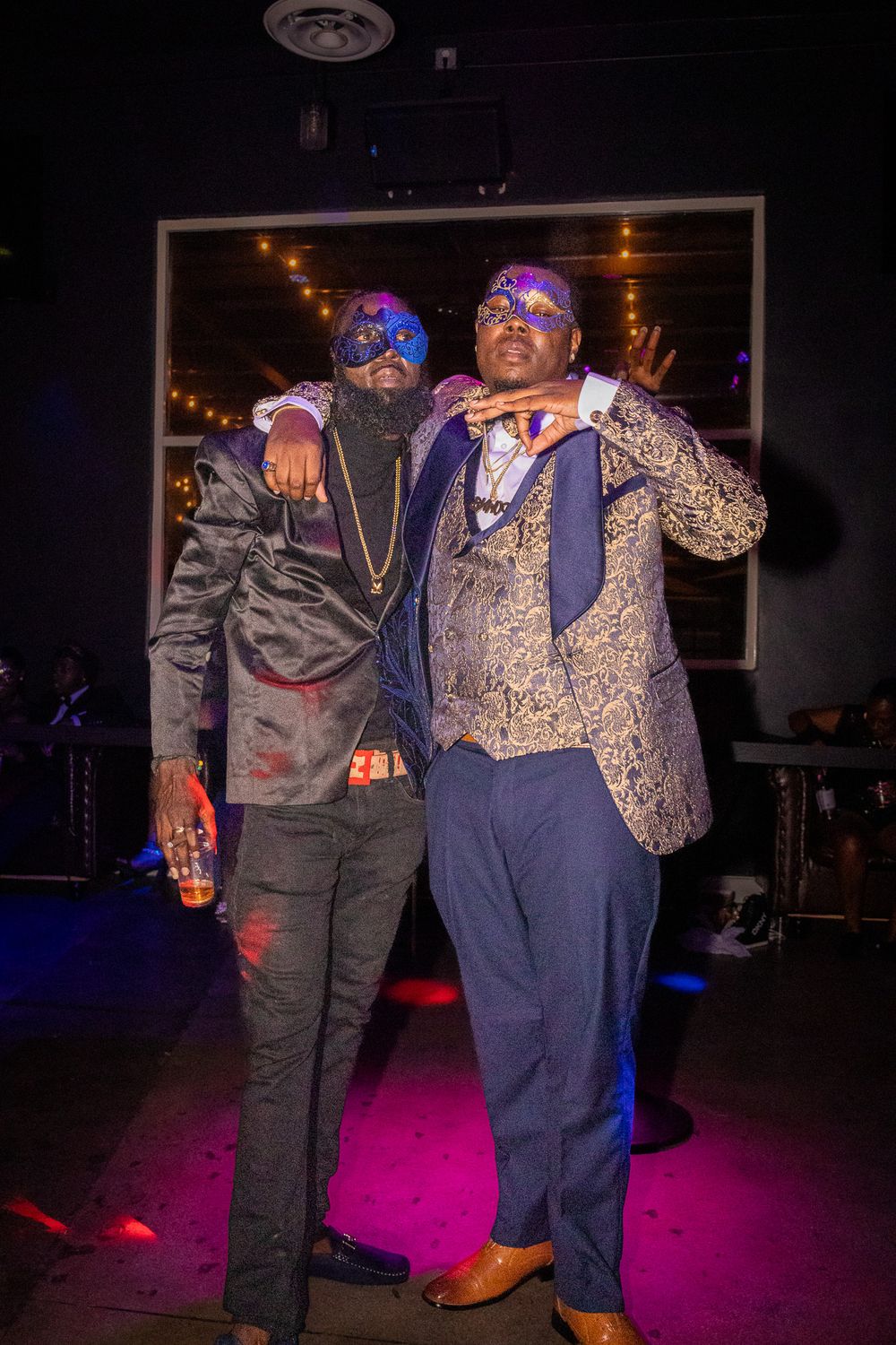 Two performers in decorated masks and formal wear pose on a nightclub's illuminated stage.