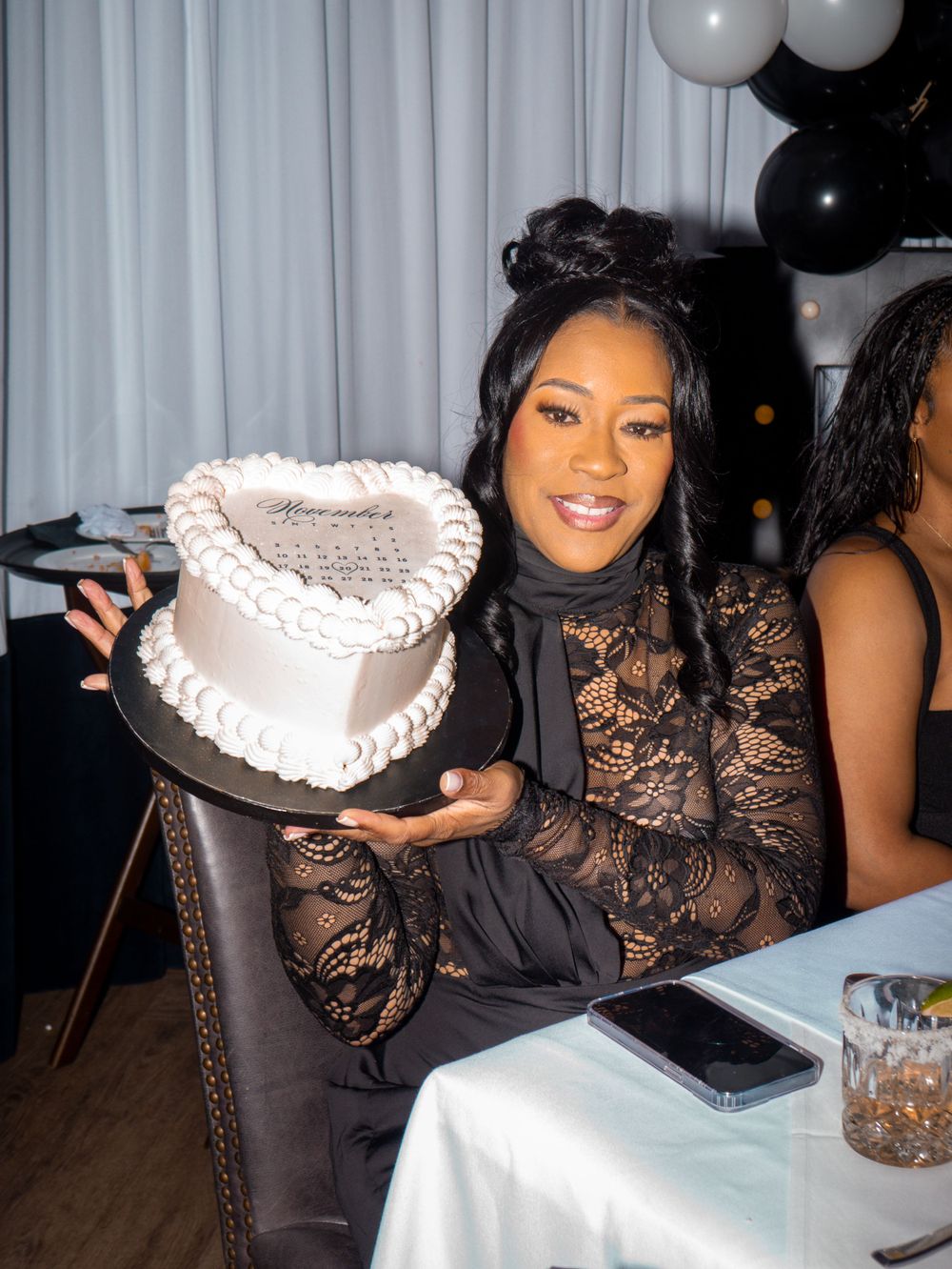 Woman in black lace dress holds decorative white cake at formal event with balloons.