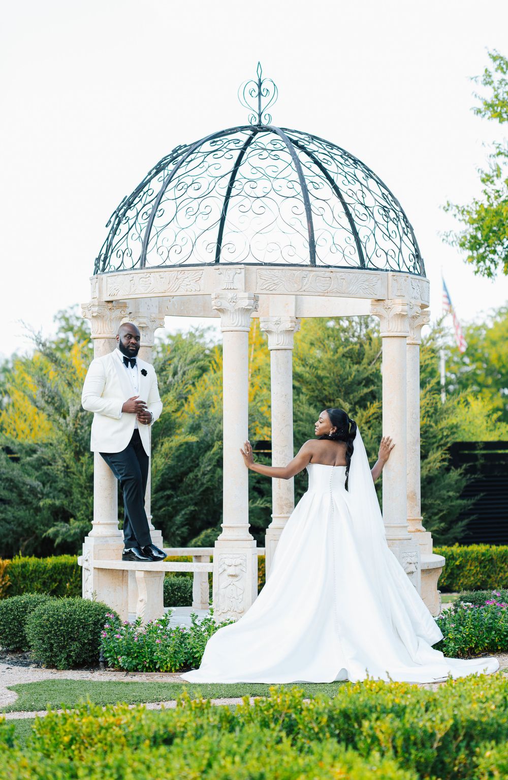 A wedding couple poses under an ornate white gazebo with blue dome in a manicured garden setting.