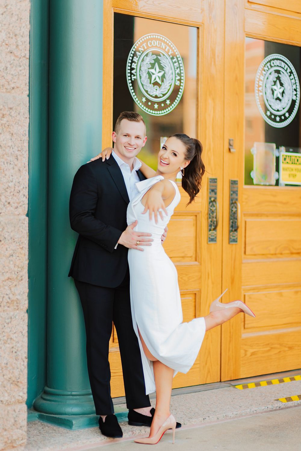 A couple in formal attire poses happily in front of wooden doors with Texas state seals at a government building.