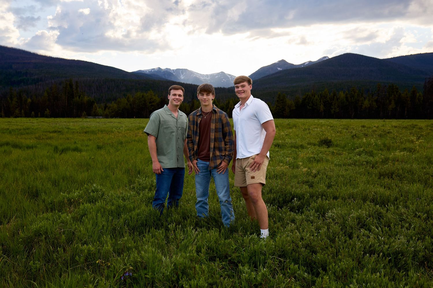 Family group standing together in a grassy meadow with majestic mountain peaks and pine trees in the background.