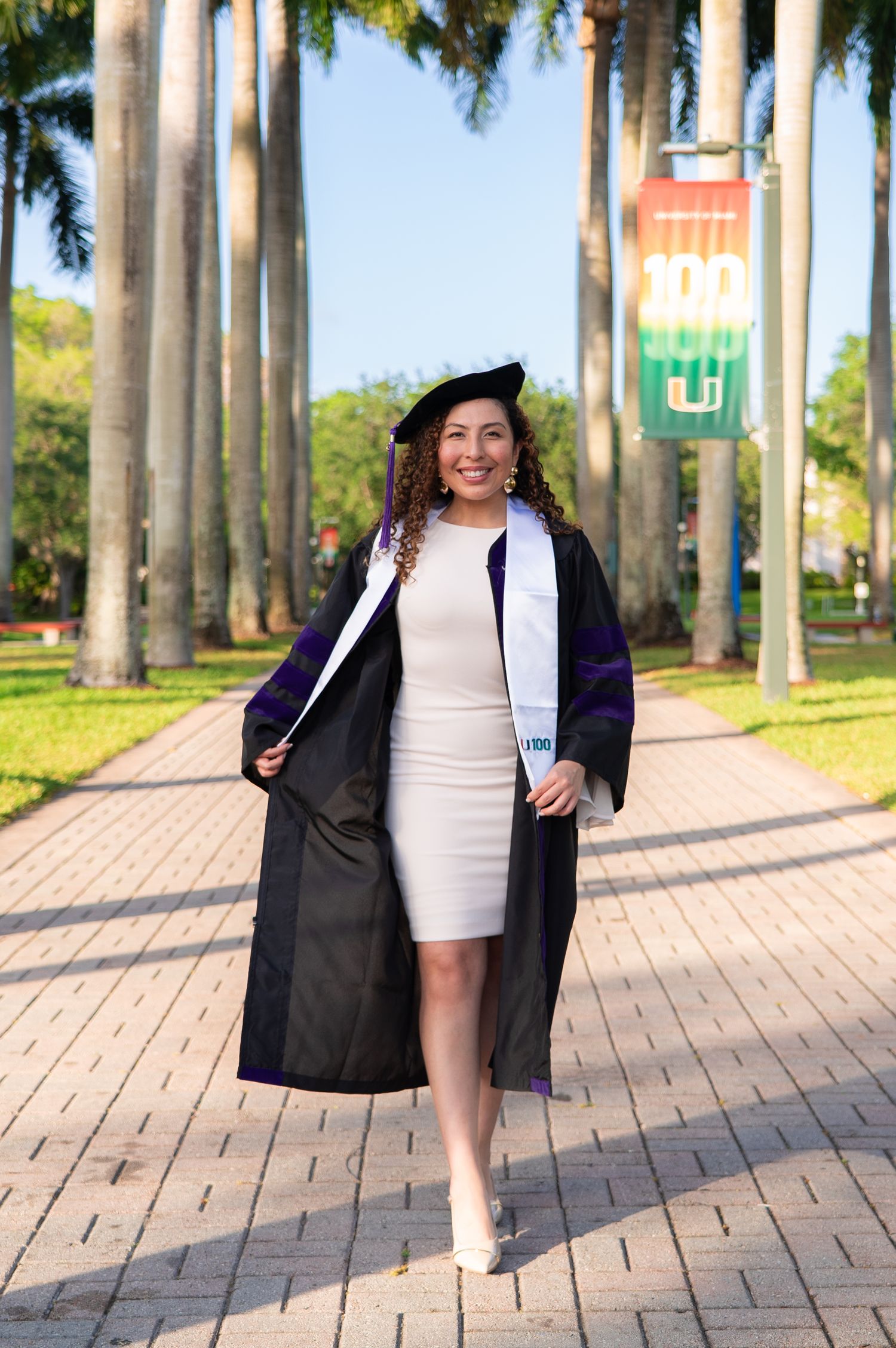 Woman in black graduation gown and cap walks on brick path at University of Miami campus.