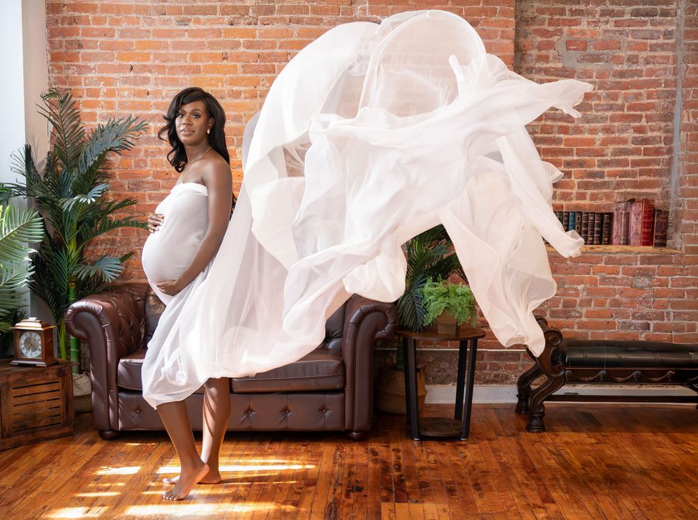 Pregnant woman in white flowing fabric gown poses elegantly in a rustic brick-walled studio during maternity photoshoot.